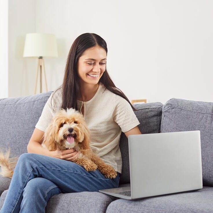 Happy Female Holding Small Dog In Lap On Couch While Using Laptop